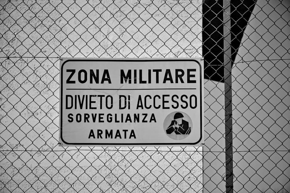 Black and white photo of a restricted military zone sign behind a metal fence. Black and white photo of a restricted military zone sign behind a metal fence.