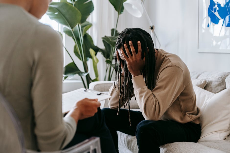 Anxious African American male with hands on head sitting on sofa near crop female psychologist with clipboard during appointment in office