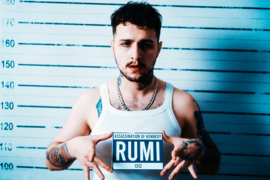 Portrait of a tattooed man holding a sign against a mugshot backdrop. Portrait of a tattooed man holding a sign against a mugshot backdrop.