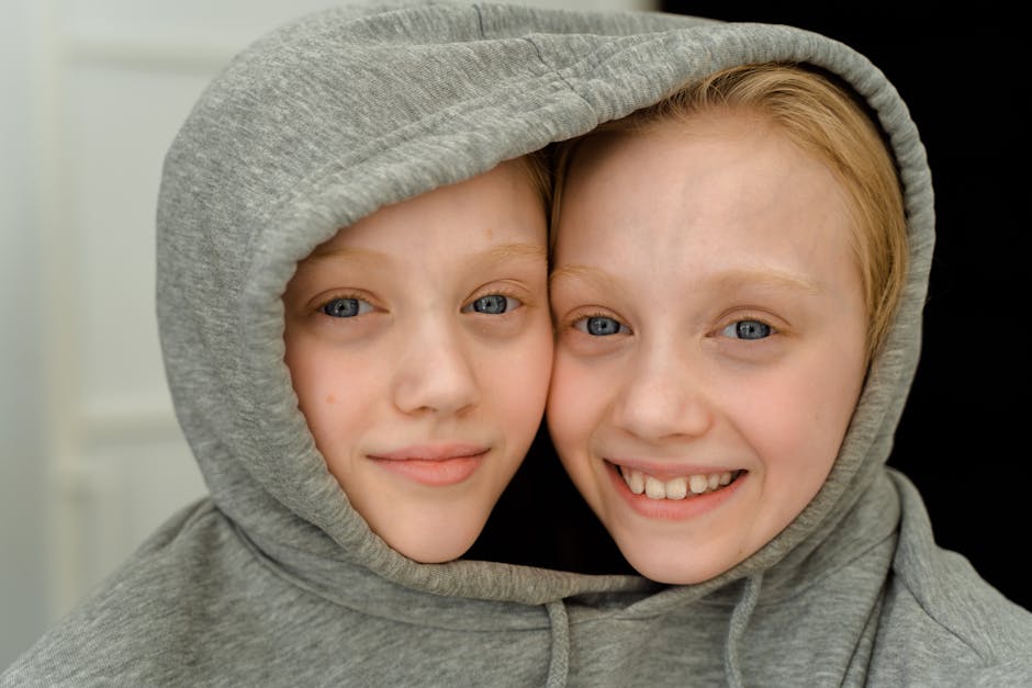 Charming close-up of smiling twin girls with blonde hair sharing a grey hoodie indoors. Charming close-up of smiling twin girls with blonde hair sharing a grey hoodie indoors.